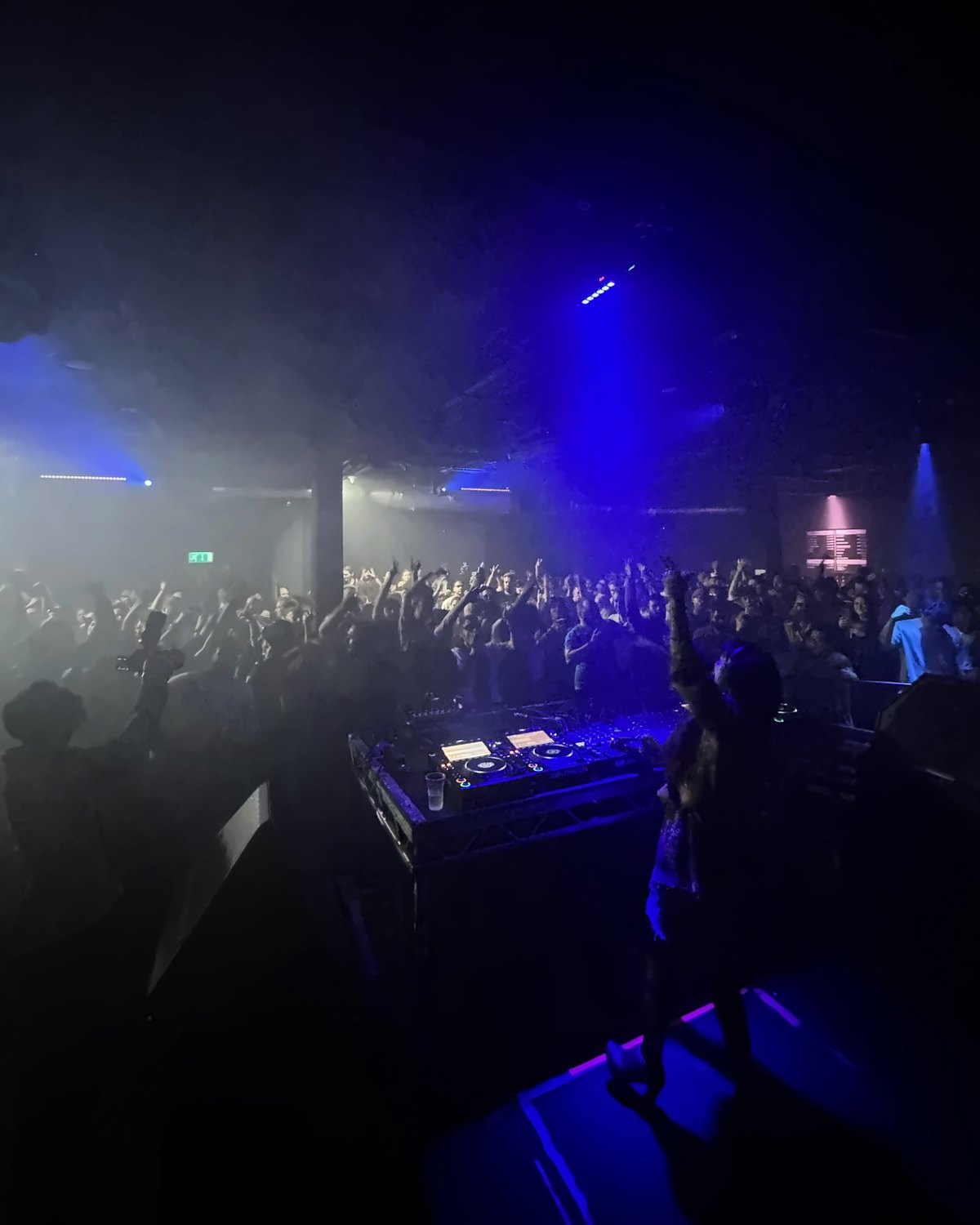 Lily Rivers behind the decks, arm raised to a full dance floor in blue haze